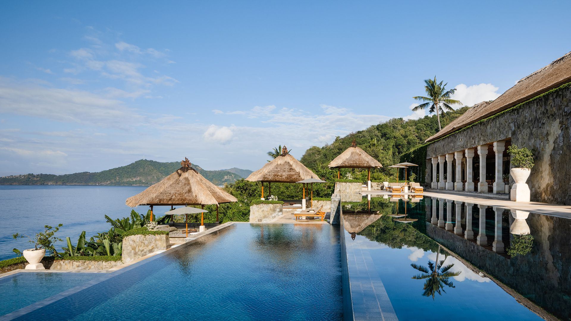 Amankila's famous three-tier infinity pool cascading toward the Lombok Strait, with thatched bales along the edge