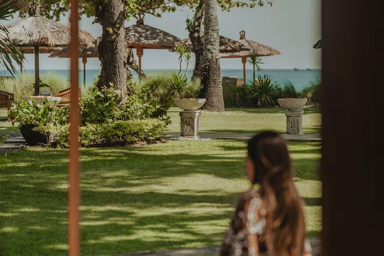 A guest looking out over Jimbaran Puri's lawn toward the sea from beneath a garden umbrella